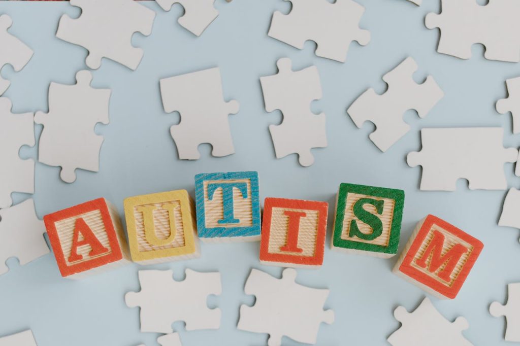 wood-letter-blocks-spelled-autism-8386122 Colorful blocks spelling autism amidst scattered puzzle pieces on a light blue background, symbolizing autism awareness.